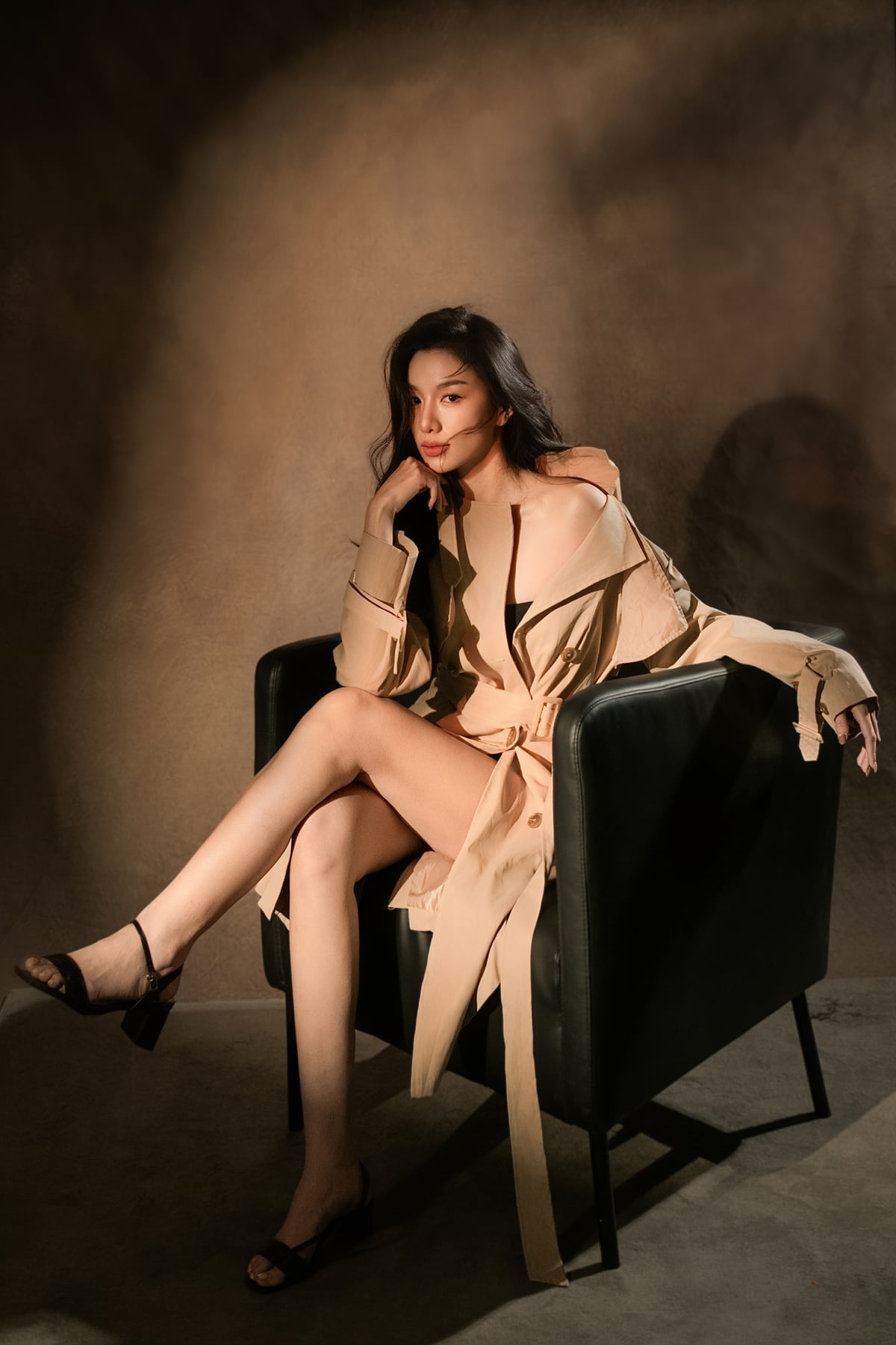 K9 Studio client portrait in a beige trench coat, seated in an armchair, moody warm-toned lighting