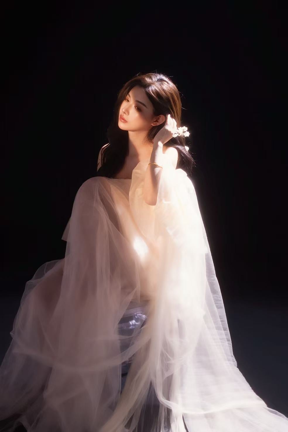K9 Studio client portrait in a white tulle dress, seated, hand raised to the hair, dark background