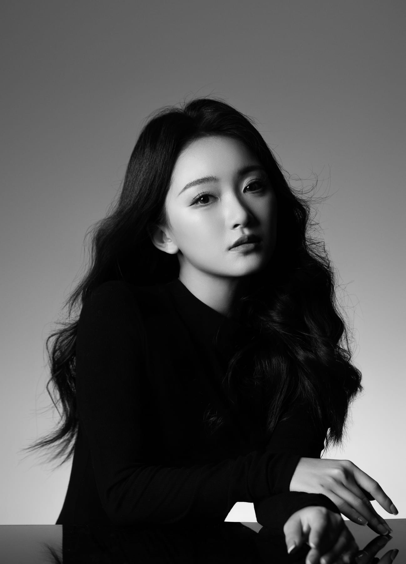 K9 Studio client black-and-white beauty portrait in a black top, seated at a reflective black surface, hand resting on the table, looking upward